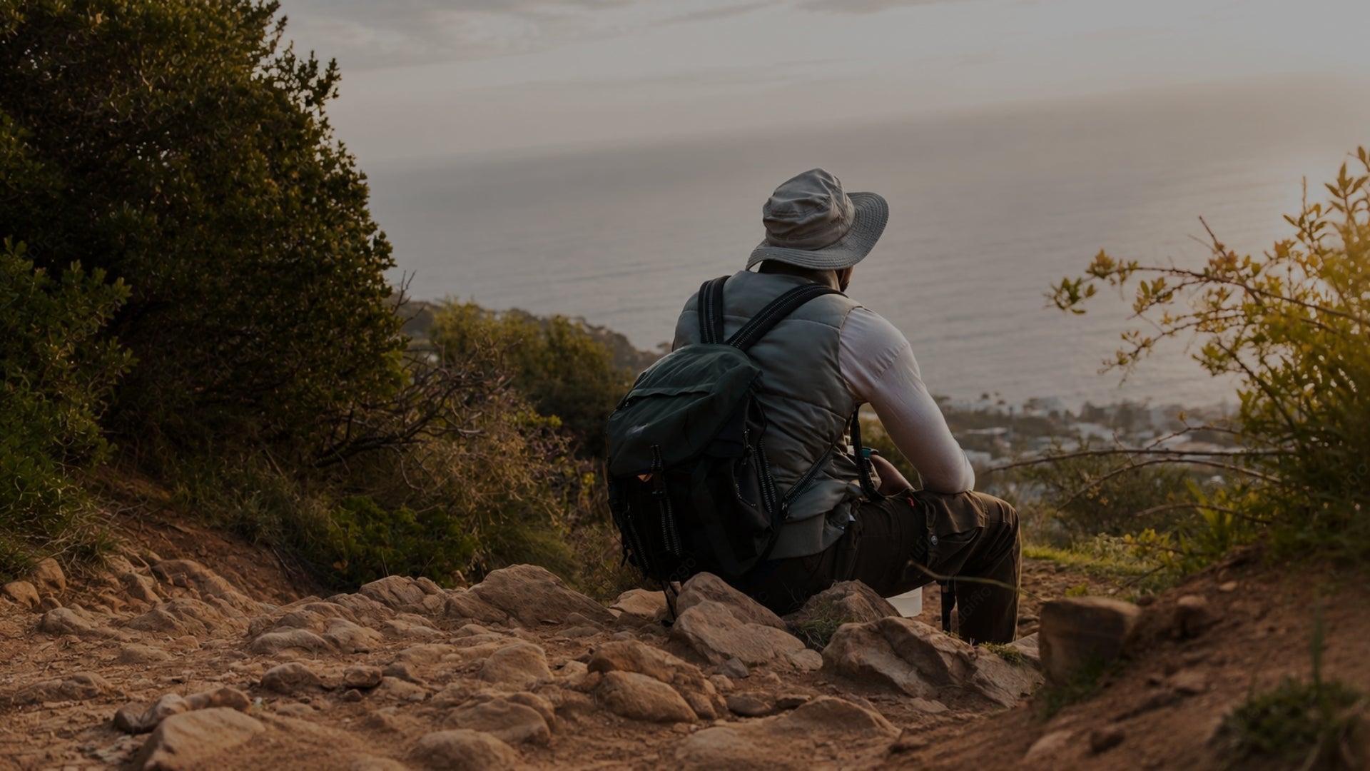 A person sitting on a rocky hillside outdoors, seen from behind, wearing a wide-brim hat, backpack, and long-sleeve shirt while looking out over a coastal town at sunset.