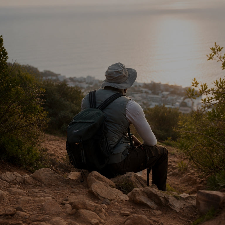 A person sitting on a rocky hillside outdoors, seen from behind, wearing a wide-brim hat, backpack, and long-sleeve shirt while looking out over a coastal town at sunset.